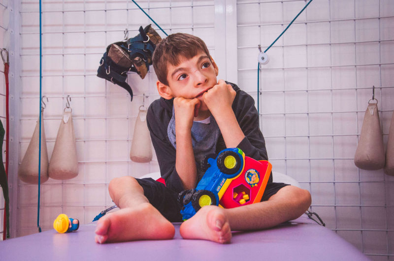 Menino com microcefalia, durante uma sessão de fisioterapia. Ele está sentado em uma maca, com as mãos no queixo e olhando para o lado.