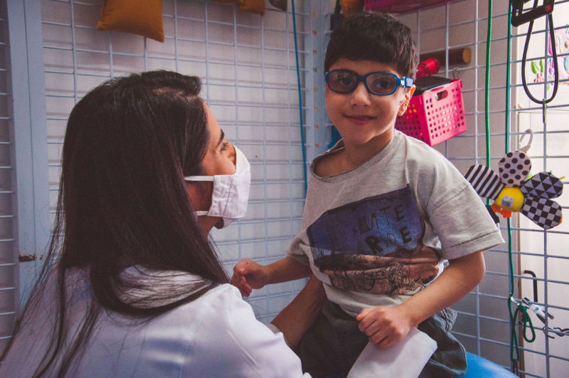 Fisioterapeuta atendendo um menino sorridente com paralisia cerebral. Ele está em cima de uma bola Bobath