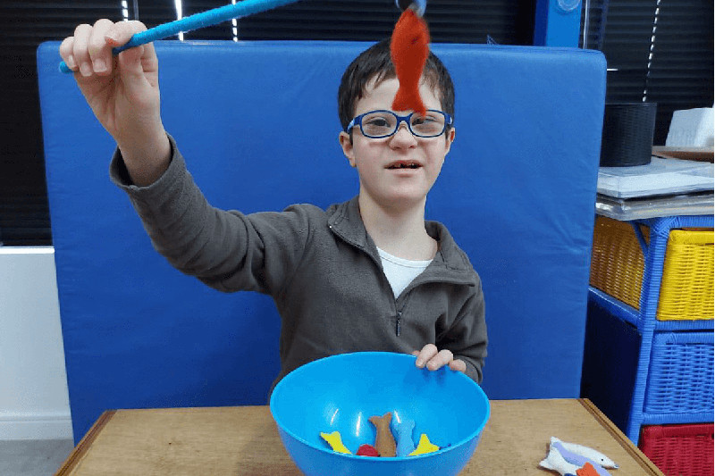 Menino com Síndrome de Down durante uma sessão de terapia ocupacional, ele está pescando um peixe de brinquedo e olhando para o peixe com um sorriso